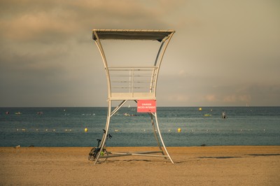 Poste de surveillance vide sur une plage de Marseille au petit matin, avec vélo, panneau rouge, sable, mer Méditerranée et ciel nuageux.