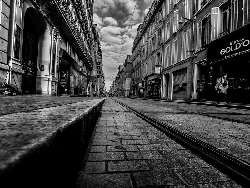 Vue au ras du sol de la rue de Rome à Marseille, avec rails du tramway, pavés, façades anciennes et commerces du centre-ville.