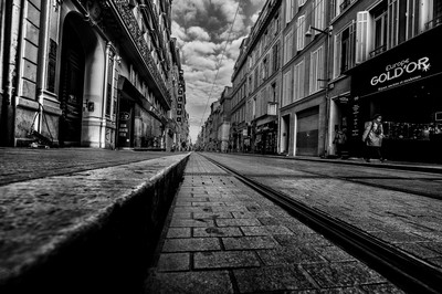 Vue au ras du sol de la rue de Rome à Marseille, avec rails du tramway, pavés, façades anciennes et commerces du centre-ville.